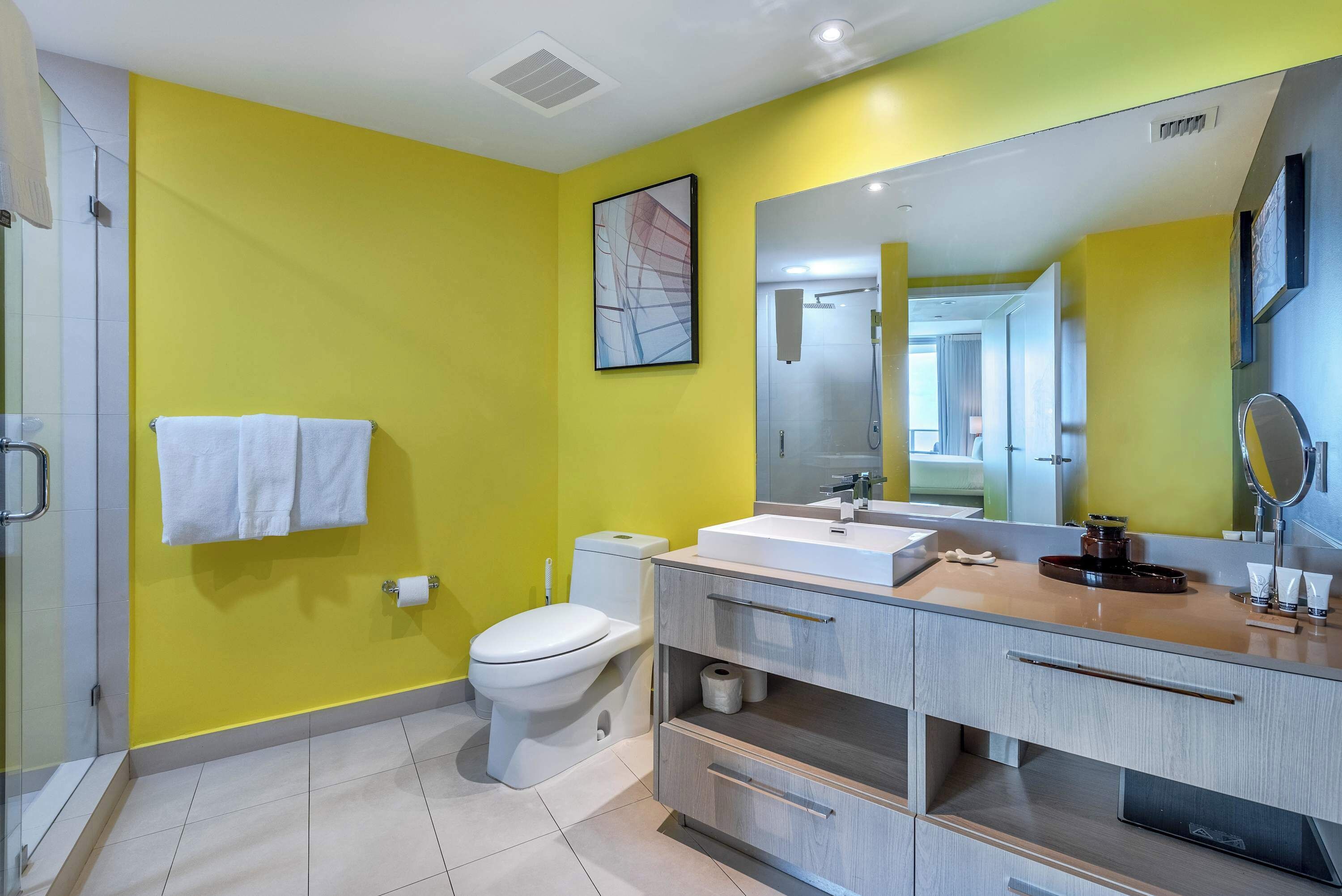 A modern bathroom with a yellow wall, a white toilet, a sink with a mirror, and a shower area.