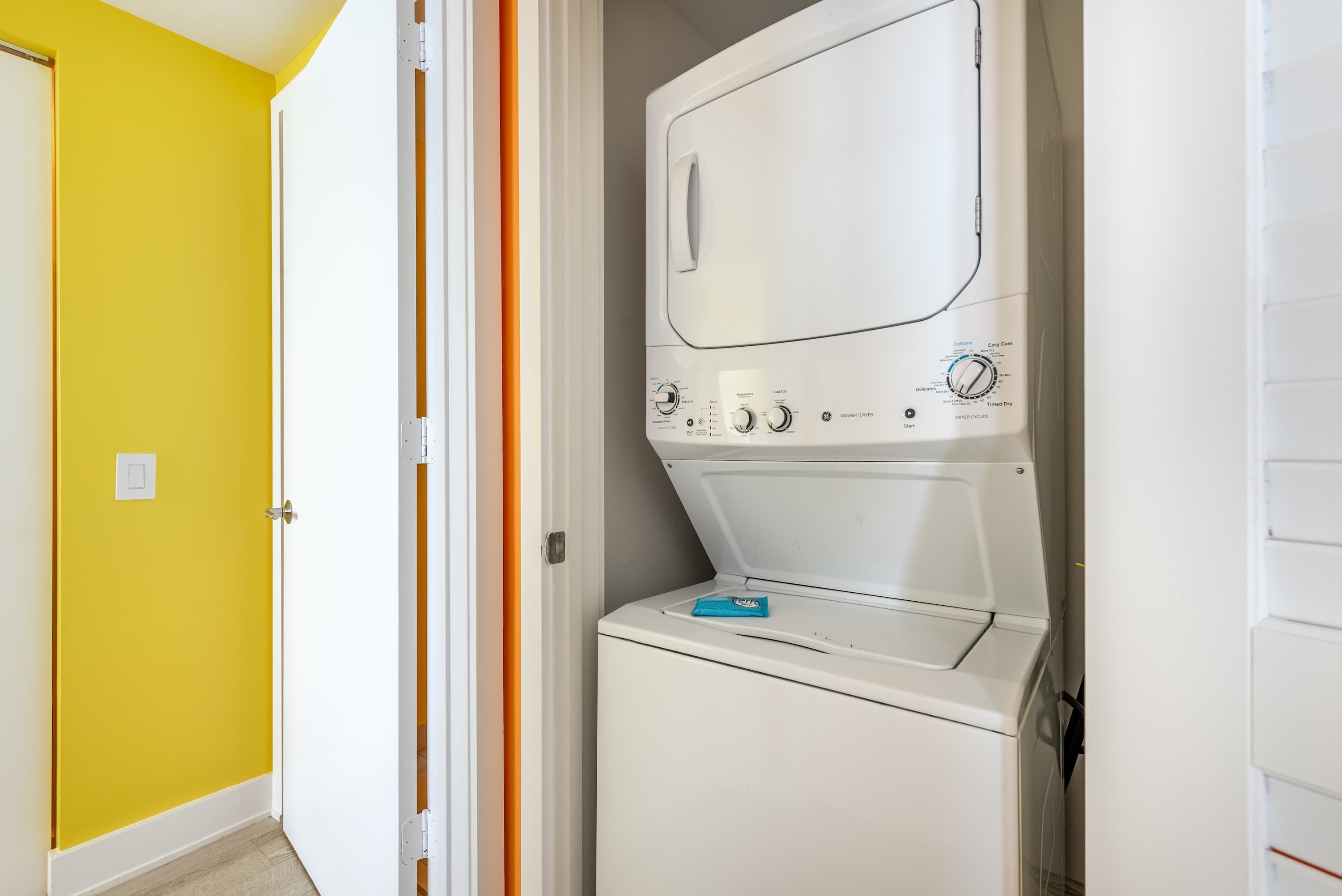 Una lavadora y secadora compactas en un lavadero pequeño con una pared amarilla y una puerta blanca.