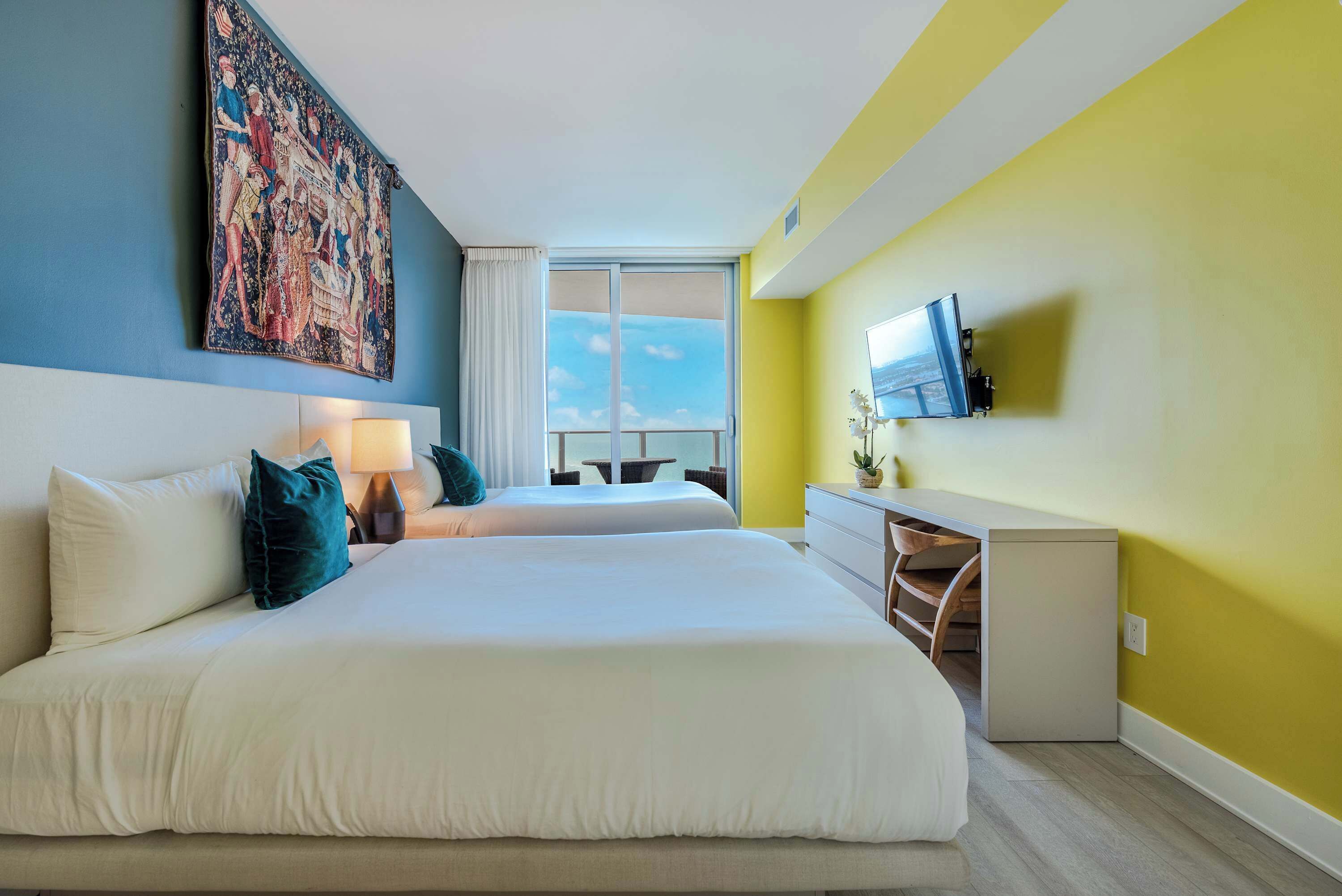 A hotel room with two beds, a large tapestry on the wall, a TV mounted on the wall, and a view of the sky through the window.