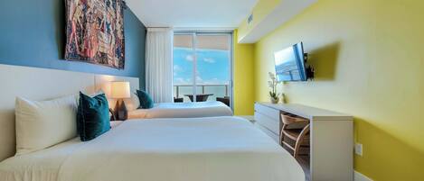 A hotel room with two beds, a large tapestry on the wall, a TV mounted on the wall, and a view of the sky through the window.