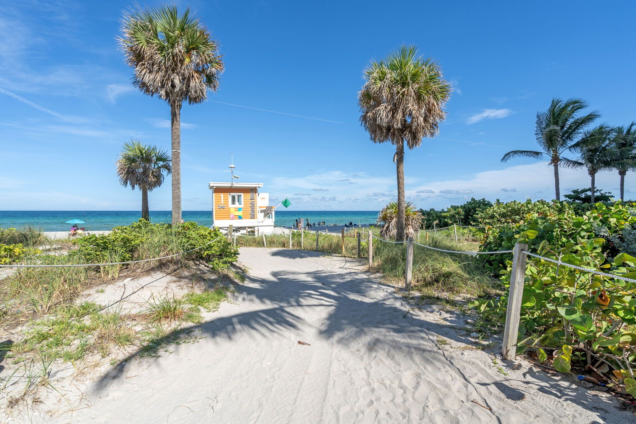 A sandy pathway leading to a beach with palm trees and a small building with a flag.