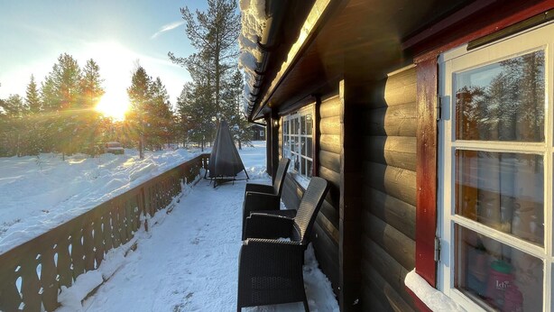 A wooden cabin with a covered porch, a window with a view of the surrounding trees, and a snow-covered ground.