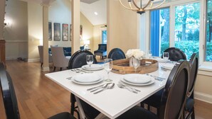 Un coin repas avec une table blanche dressée pour quatre personnes, entourée de chaises sombres, d’un lustre et offrant une vue sur la verdure à l’extérieur.