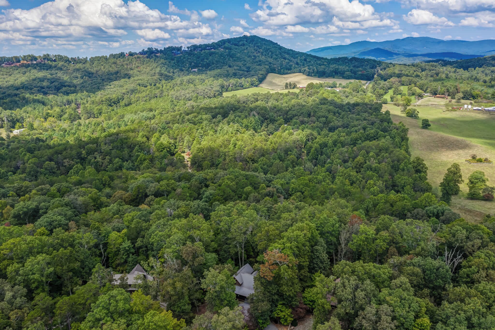 A lush, green landscape with a house nestled among dense trees, surrounded by rolling hills and distant mountains.