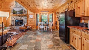 Interior de una cabaña de troncos con chimenea de piedra, una mesa de comedor de madera y una zona de cocina con refrigerador.
