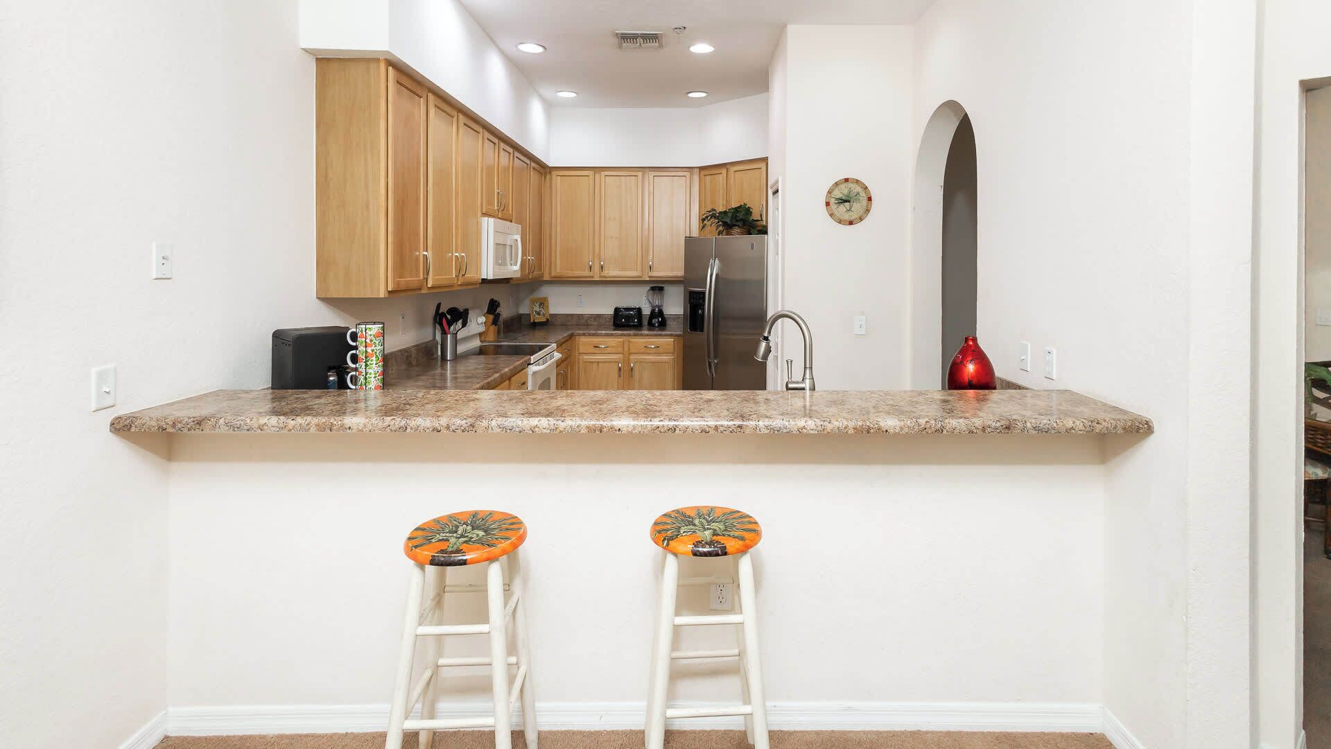 A kitchen with wooden cabinets, a stainless steel refrigerator, a microwave, and a sink. There are two stools with orange and green patterned seats.