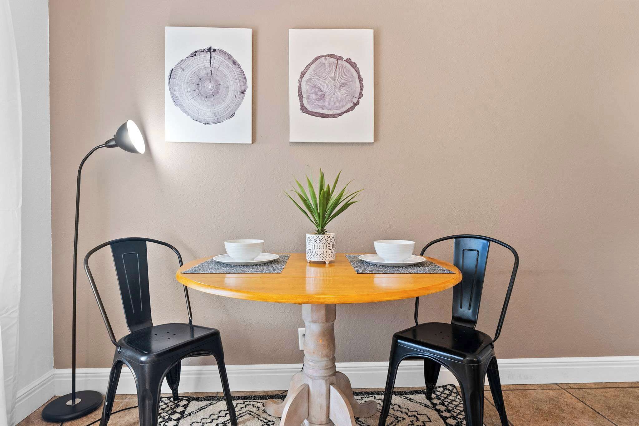 A round wooden table with two black chairs, a potted plant, and two white bowls on a patterned rug.