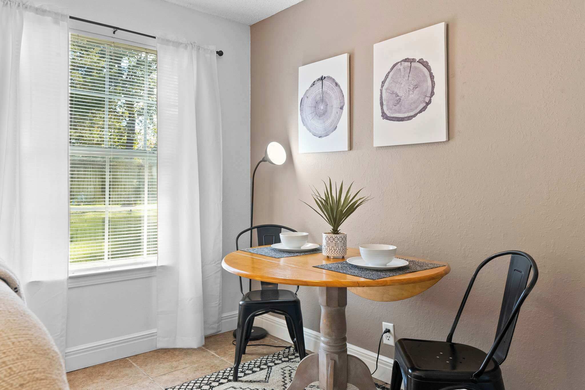 Ein Essbereich mit einem runden Holztisch, zwei schwarzen Stühlen, einem Blumentopf und zwei gerahmten Kunstwerken mit Holzmaserung an der Wand.