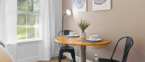 Ein Essbereich mit einem runden Holztisch, zwei schwarzen Stühlen, einem Blumentopf und zwei gerahmten Kunstwerken mit Holzmaserung an der Wand.