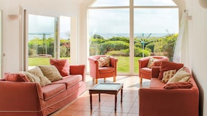 Un salon spacieux doté d’une grande porte vitrée donnant sur un jardin, de deux canapés rouges, d’une table basse en bois et d’une vue sur la mer.