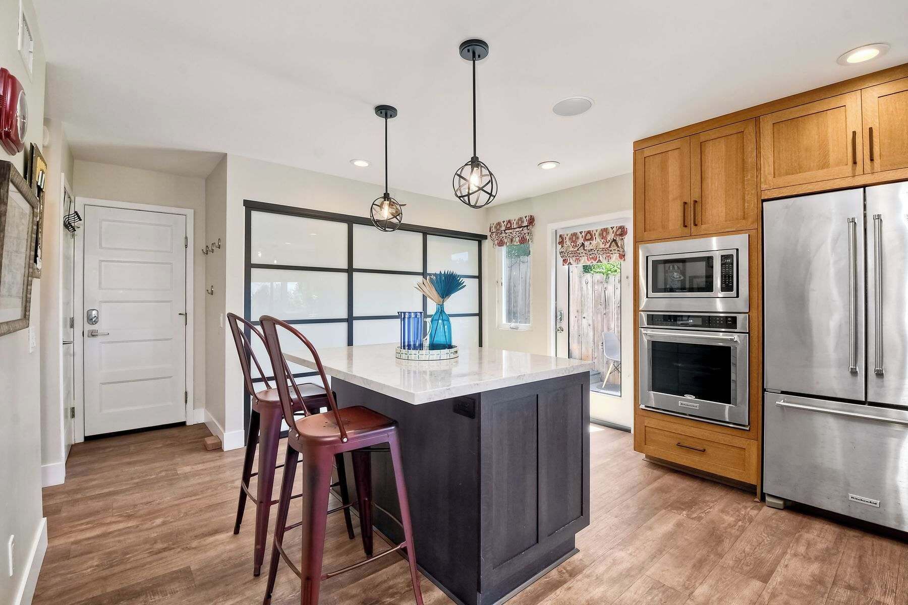 A modern kitchen with wooden cabinets, a stainless steel refrigerator, a double oven, and a bar stool.