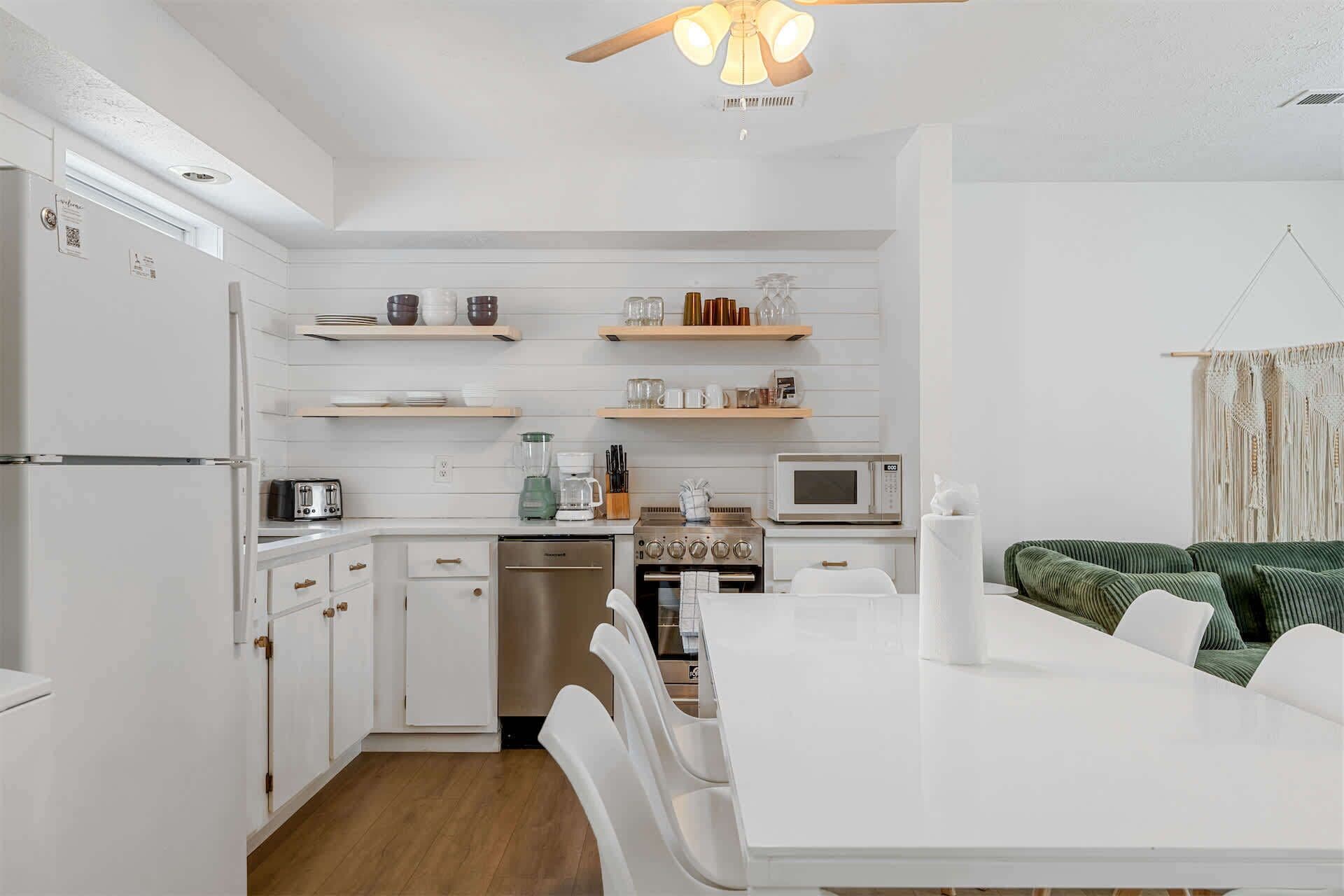 Cocina compacta con muebles blancos, lavavajillas, estufa y microondas. Hay repisas de madera sobre la encimera con varios utensilios de cocina.