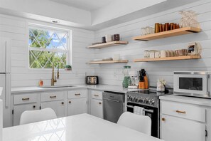 A modern kitchen with white cabinetry, a stainless steel refrigerator, a microwave, a stove, and a dishwasher. There are wooden shelves above the counter holding various kitchen items. - Tranquil Woods Retreat Branson (Branson)