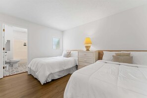Une chambre avec deux lits, une table de chevet avec une lampe, une fenêtre et une porte donnant sur une salle de bain.