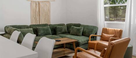 Una sala de estar con un sofá verde, una mesa de centro de madera, un sillón de cuero marrón y una mesa blanca.