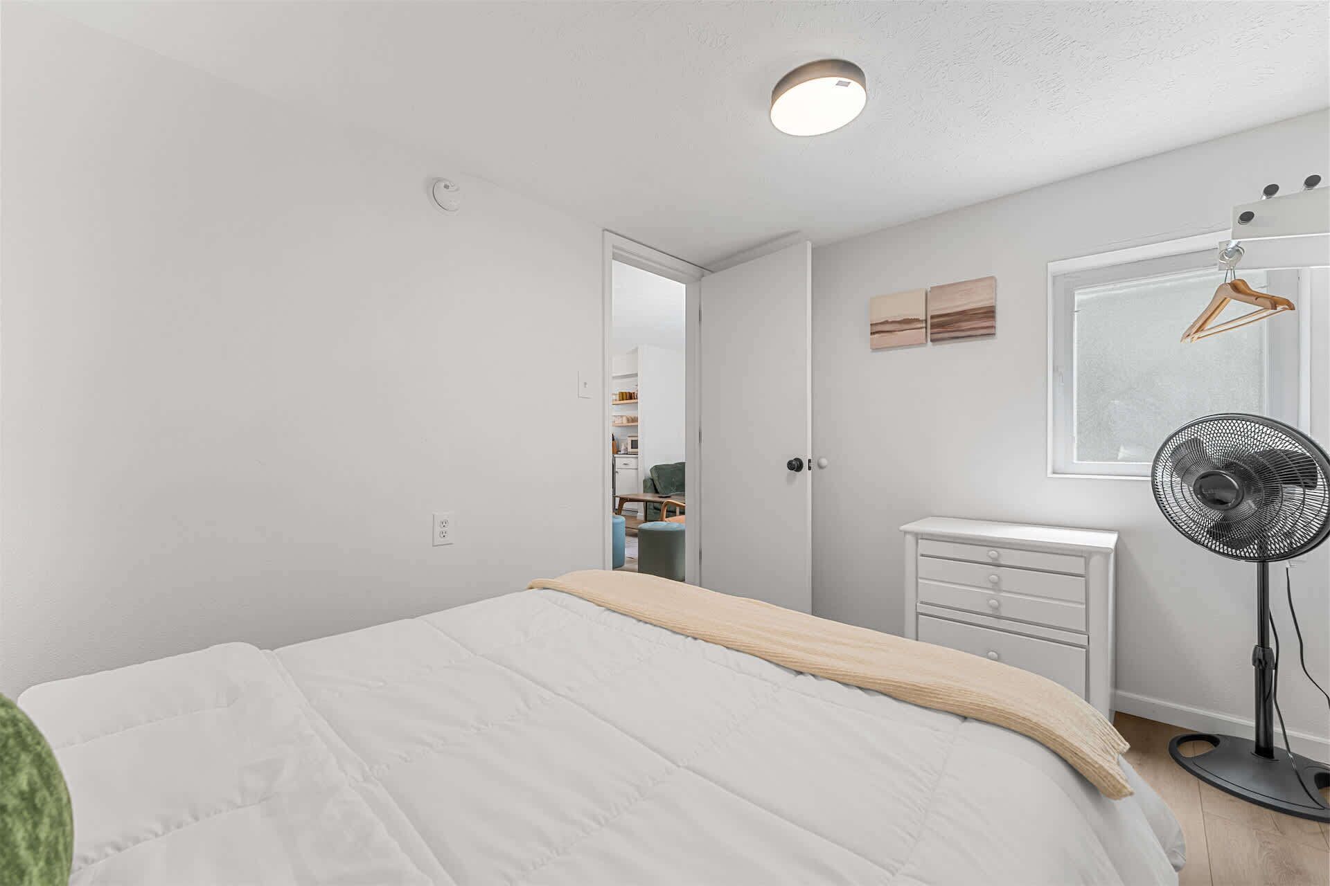 A bedroom with a bed, a dresser, a fan, and a window with a view.