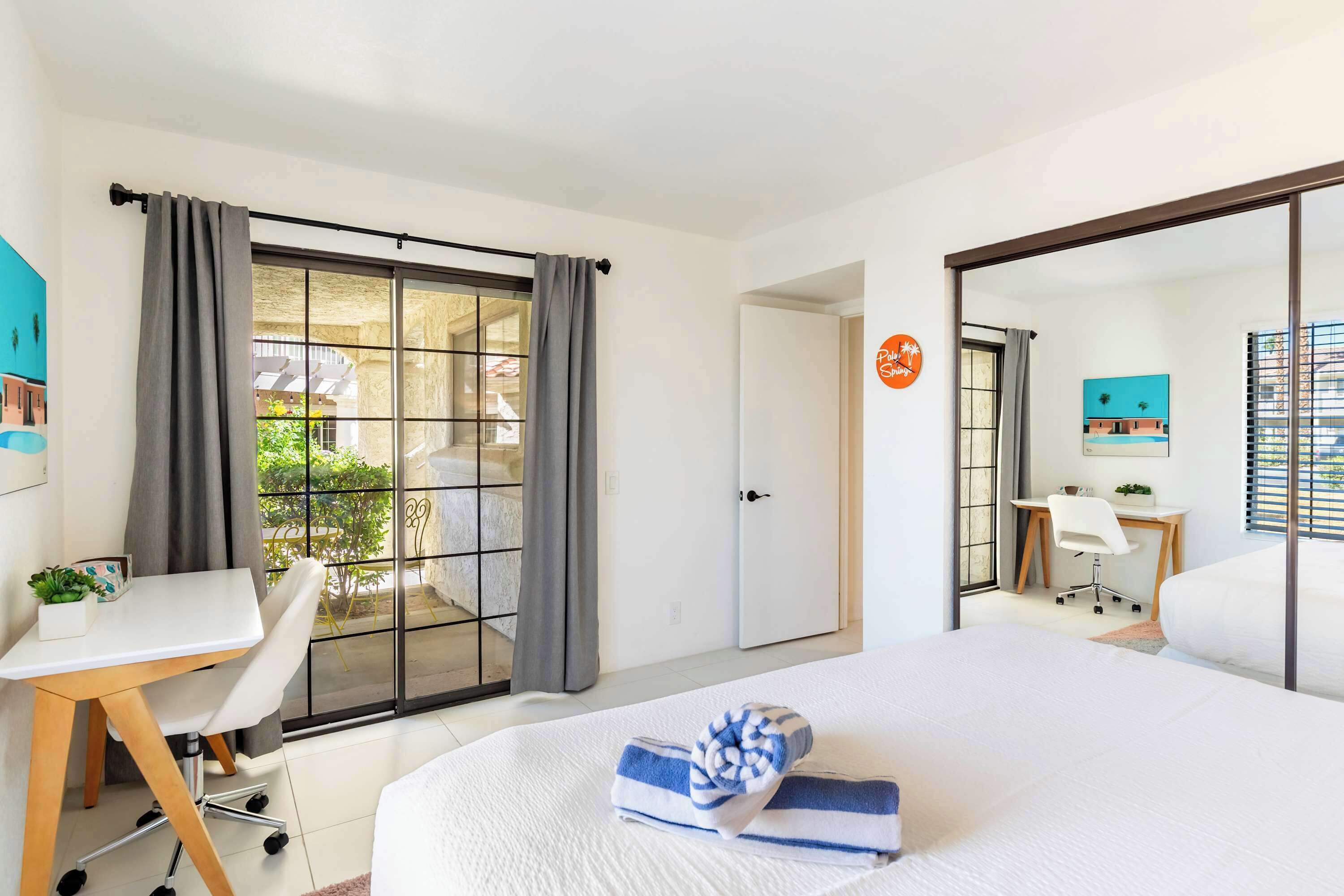A modern hotel room with a large bed, a desk with a chair, a small table with a plant, and a sliding door leading to a balcony.