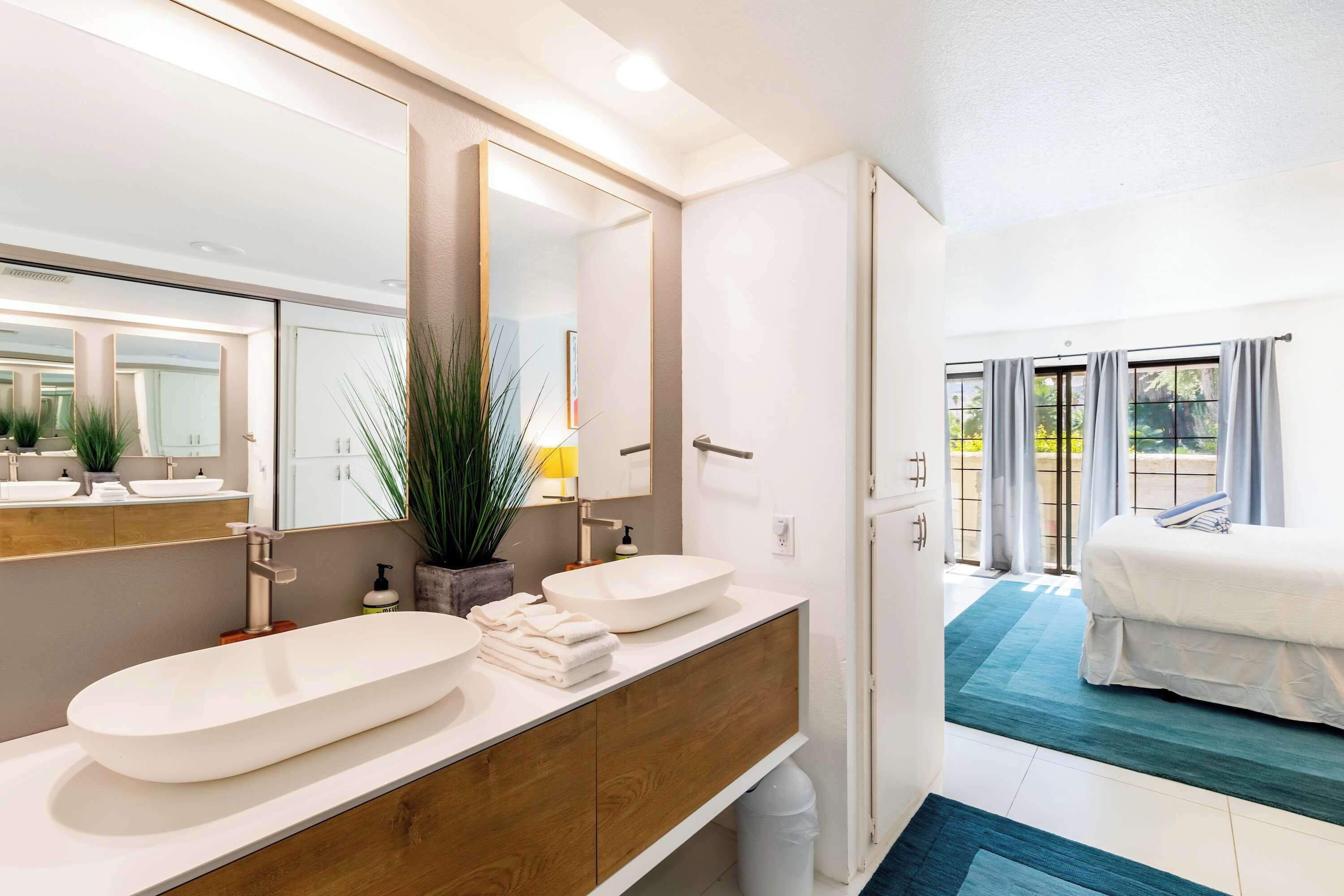Ein modernes Badezimmer mit zwei Waschbecken, einem großen Spiegel und Blick auf ein Schlafzimmer.