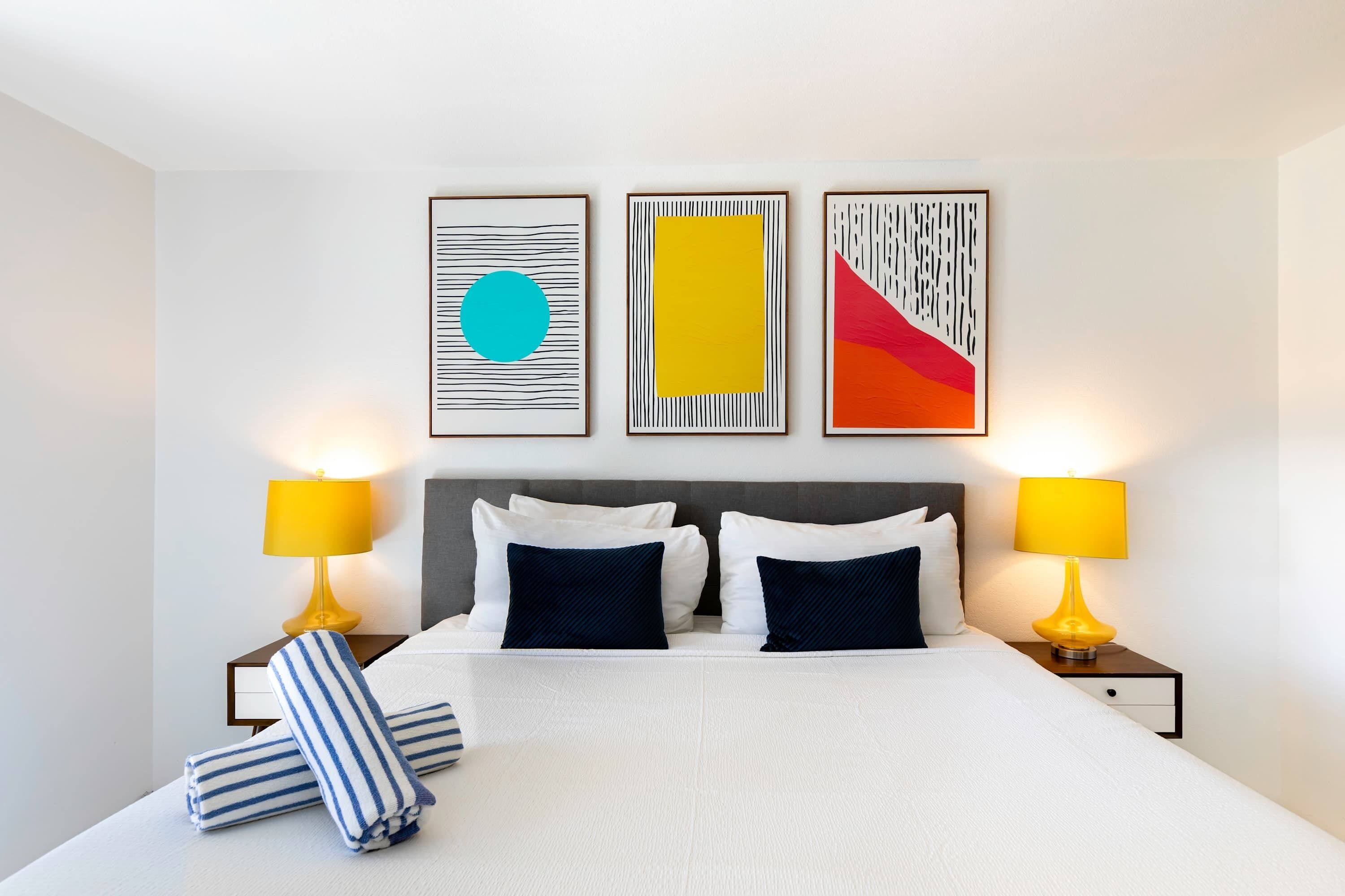 A neatly made bed with white bedding and two pillows, flanked by two bedside tables with lamps, and three framed artworks on the wall.