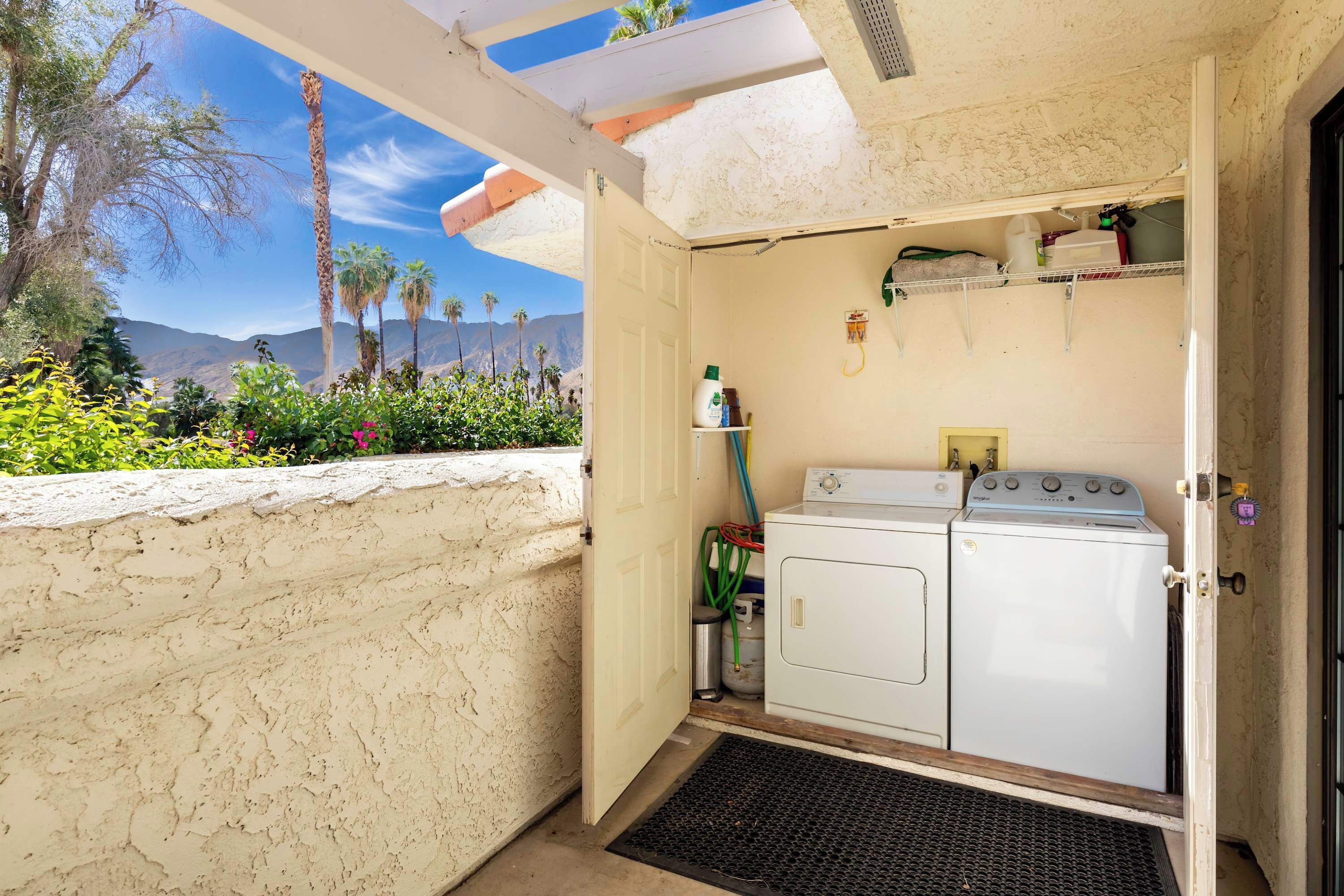 A laundry room with a washing machine and dryer, a small shelf with toiletries, and a view of palm trees and mountains.