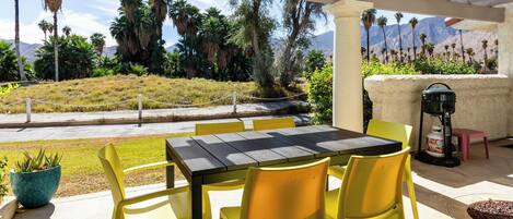 An outdoor patio with a table and yellow chairs, surrounded by palm trees and a view of mountains.