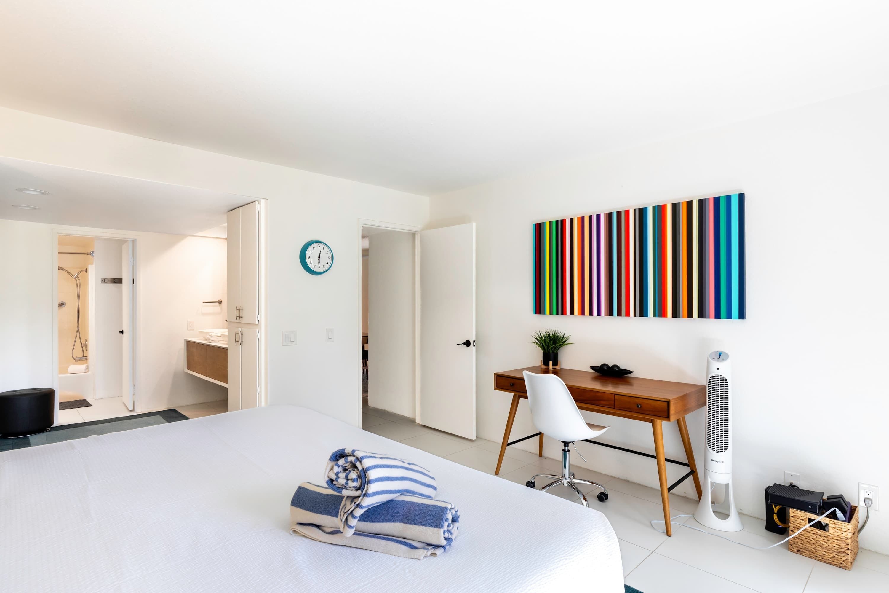 A hotel room with a bed, a desk, a chair, a standing fan, and a colorful artwork on the wall.