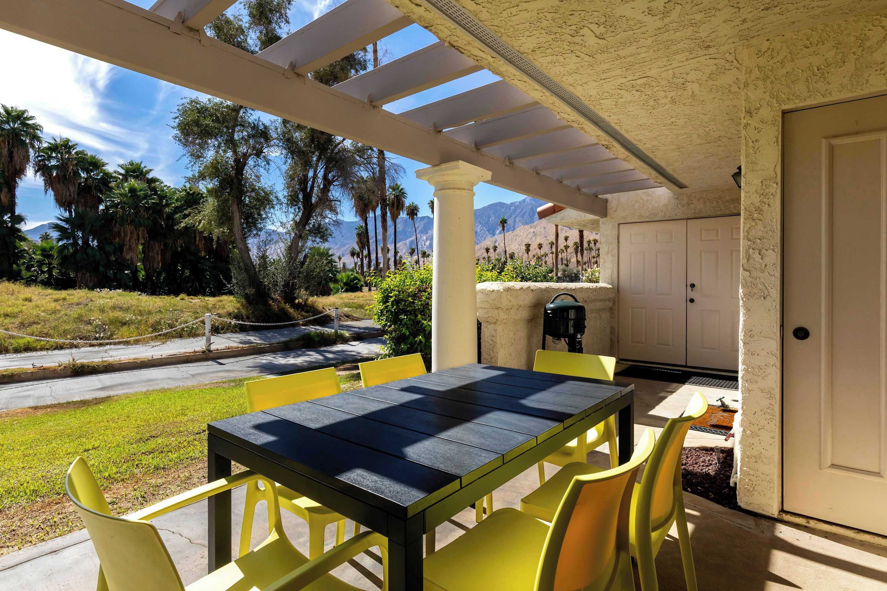 An outdoor patio with a table and chairs, a pergola, and a view of palm trees and mountains.