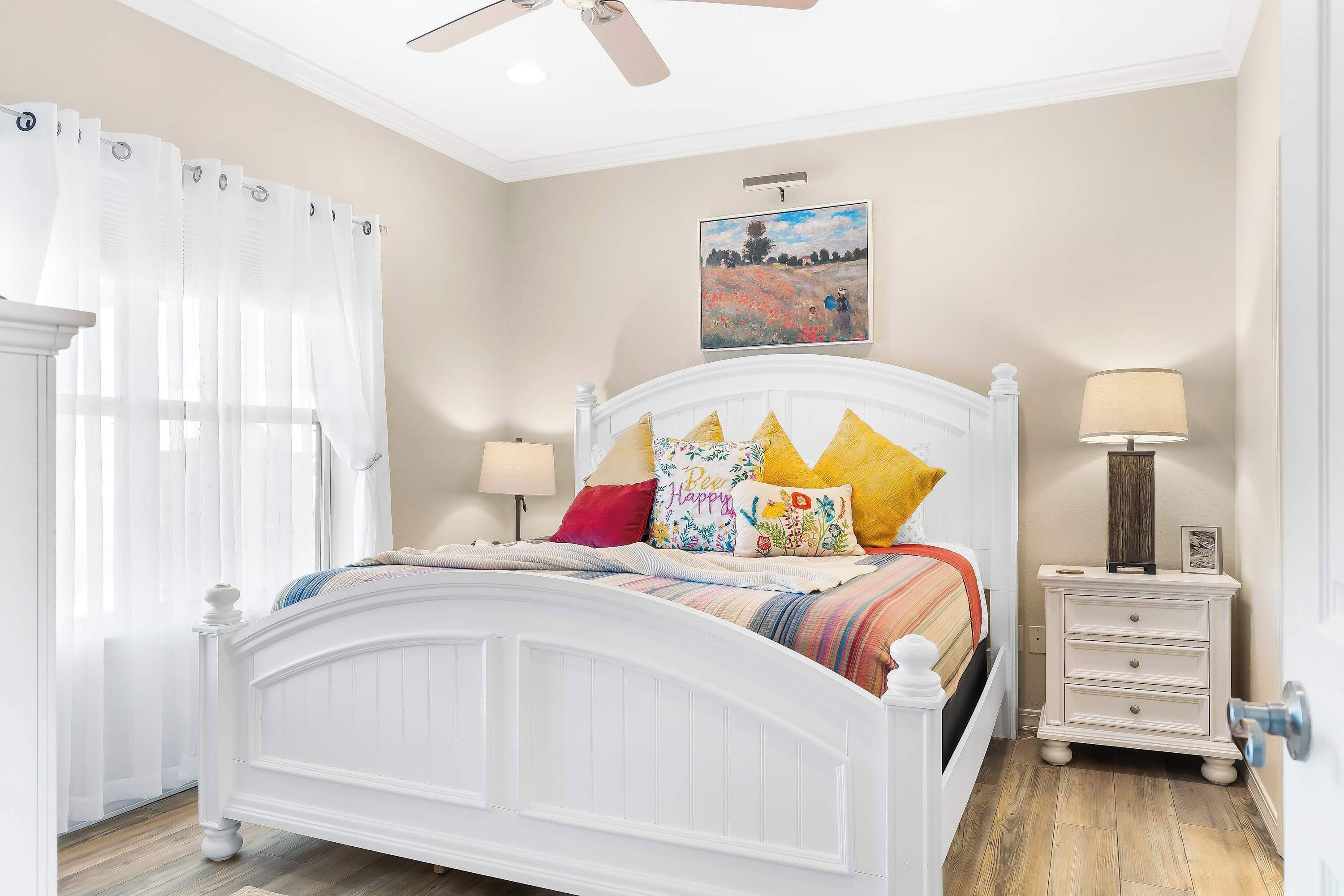 A bedroom with a white bed, colorful pillows, a nightstand with a lamp, and a painting on the wall.