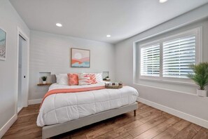 A bedroom with a bed, a window with blinds, a bedside table, and a wall-mounted shelf.