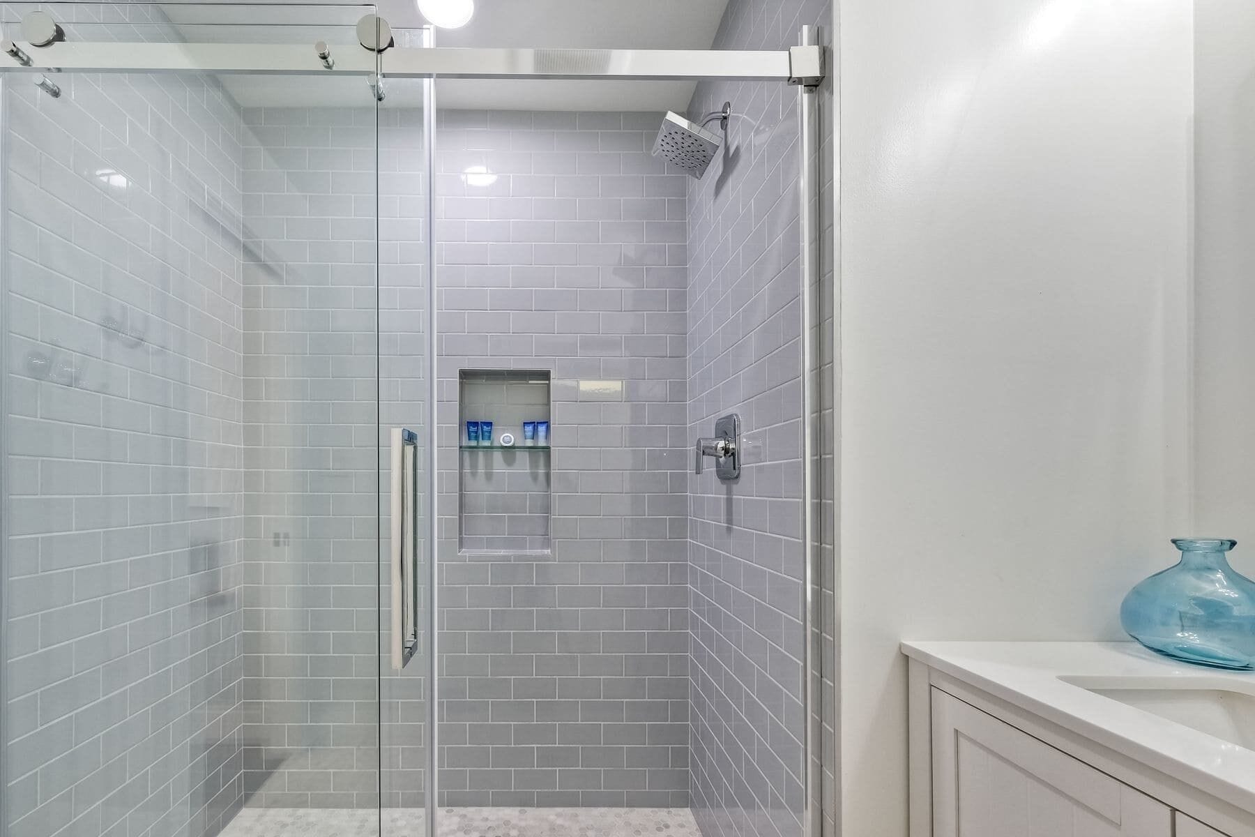 A modern shower with a glass door, grey tiled walls, and a small shelf with toiletries.
