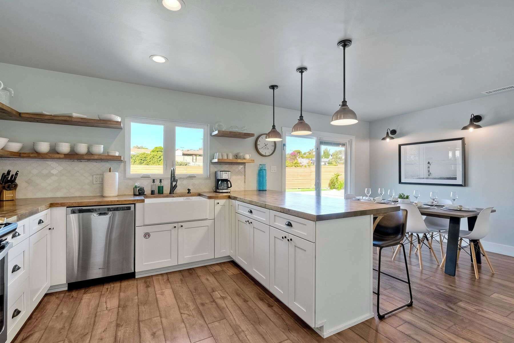 Una cocina moderna con armarios blancos, electrodomésticos de acero inoxidable y encimera de madera. Hay luces colgantes, un comedor con mesa y sillas, y vistas al exterior a través de amplias ventanas.