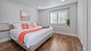 Un dormitorio con cama, ventana con persianas, una planta y un cuadro en la pared.