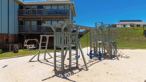 A playground with swings and climbing structures, a wooden fence, and a building with a balcony in the background. - Resort Pool & Clear Ocean Views (Port Aransas)