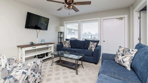 A living room with a ceiling fan, a TV mounted on the wall, a blue sofa, a glass coffee table, and a patterned armchair. - Resort Pool & Clear Ocean Views (Port Aransas)