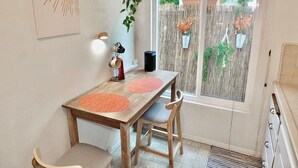 Un comedor con mesa de madera, dos sillas, estación de café y vista a través de una ventana con celosía de bambú.