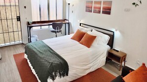Un dormitorio con una cama, un escritorio y una silla. Hay una ventana con persianas, una planta y una obra de arte en la pared.