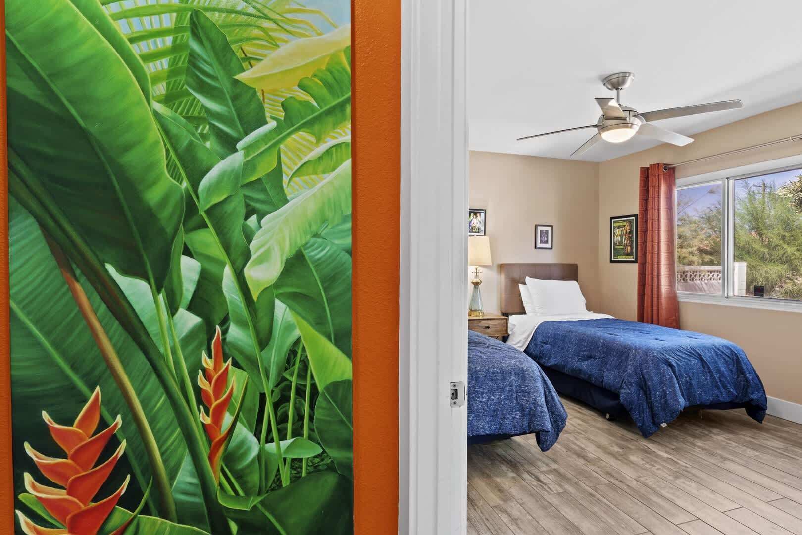 A hotel room with two beds, a ceiling fan, a window with a view, and a door with a tropical mural.