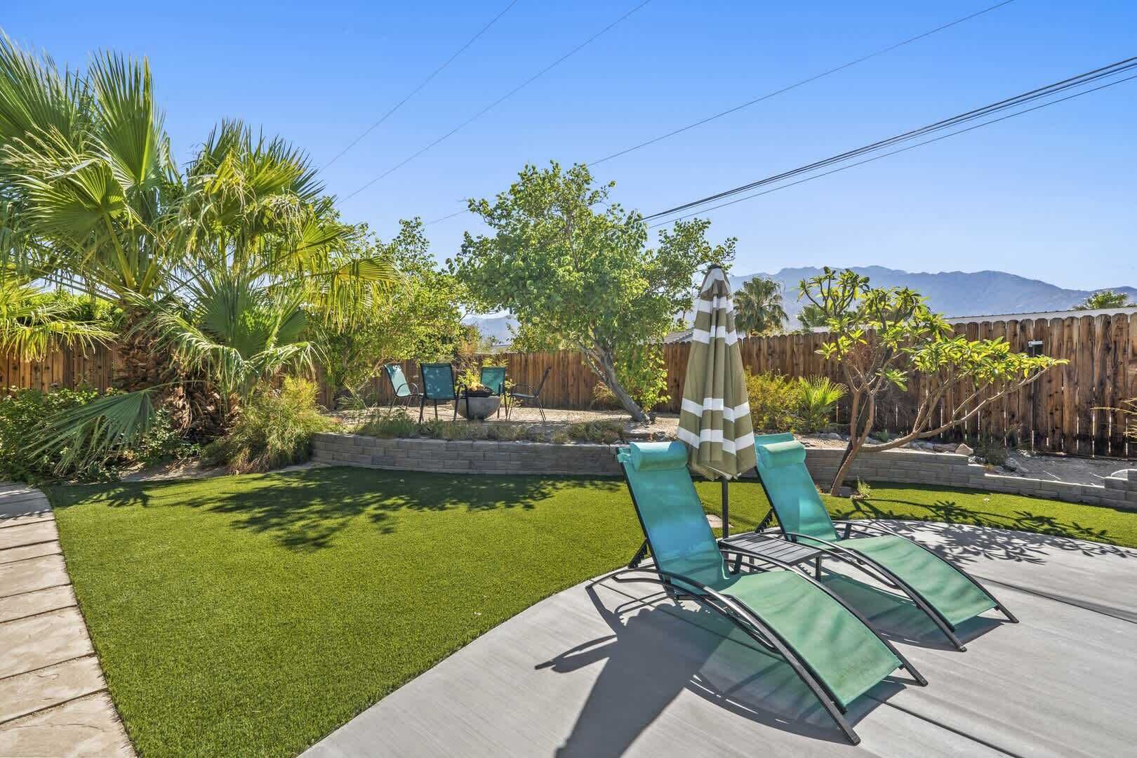 A backyard with artificial grass, two green lounge chairs, a striped umbrella, and a wooden fence.