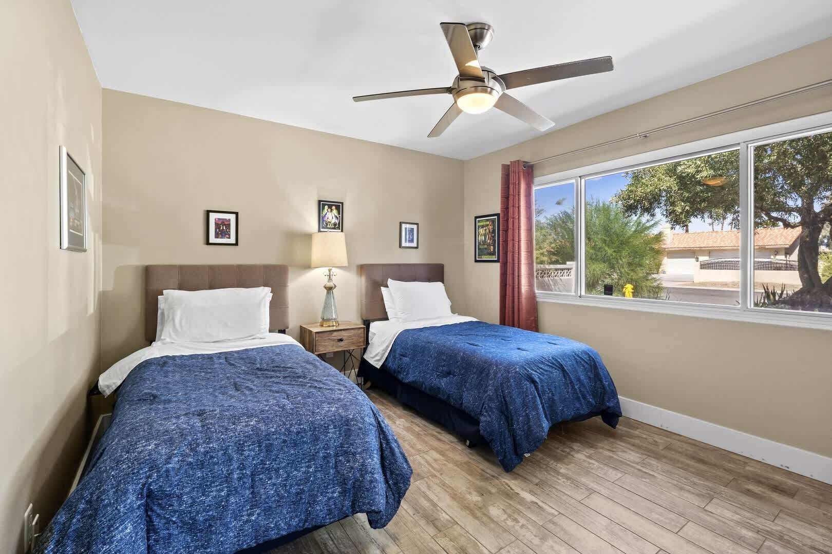 Une chambre d’hôtel avec deux lits, un ventilateur de plafond, une fenêtre donnant sur l’extérieur et des tableaux encadrés accrochés au mur.
