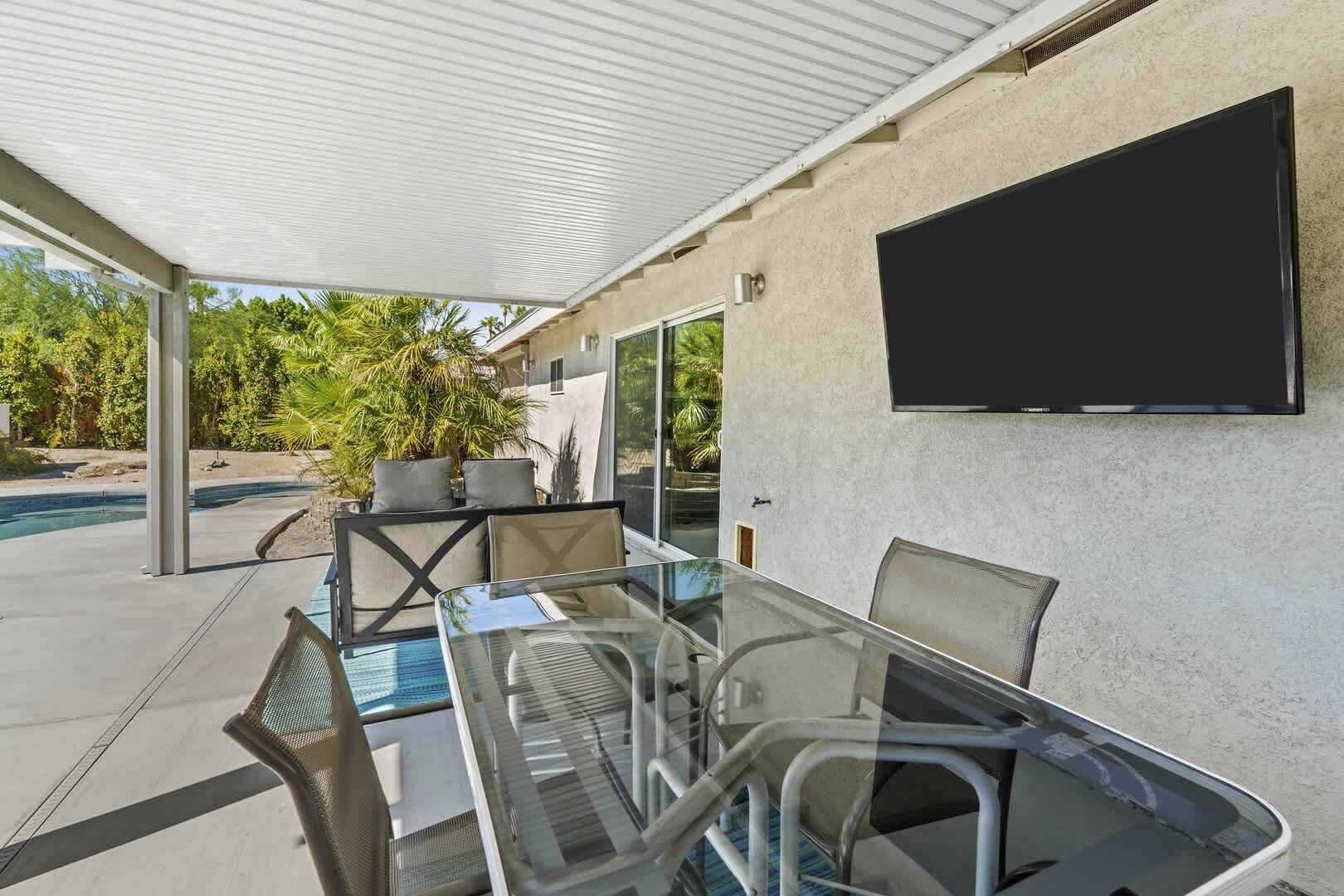 A covered outdoor area with a glass table and chairs, a flat-screen TV mounted on the wall, and a pool in the background.