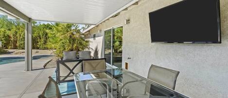 A covered outdoor area with a glass table and chairs, a flat-screen TV mounted on the wall, and a pool in the background.