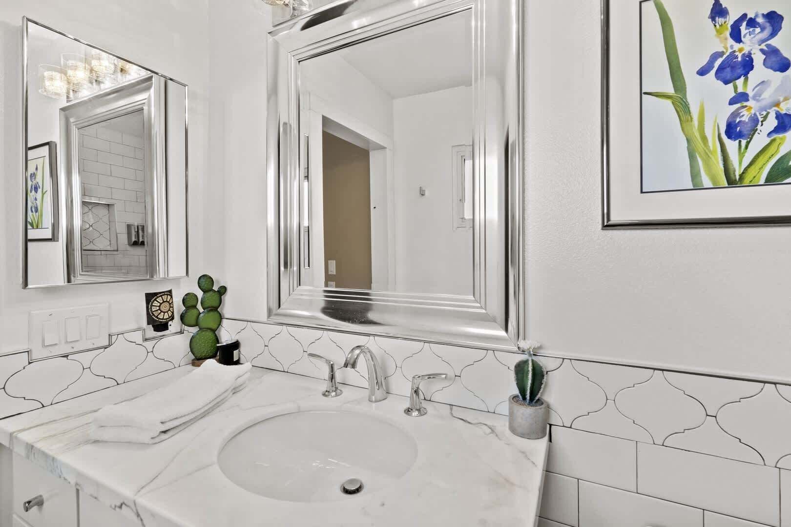 A bathroom with a white sink, two mirrors, a framed picture of blue flowers, and a cactus on the counter.