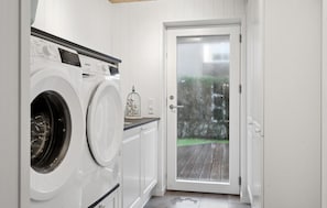 A laundry room with two front-loading washing machines, a small table with a decorative item, and a glass door leading to an outdoor wooden deck.