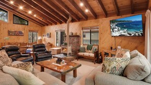 A cozy cabin interior with wooden beams, a stone fireplace, leather sofas, and a large window overlooking a scenic landscape. - Lake Tahoe Treasure! Minutes From Beach & Skiing (Homewood)