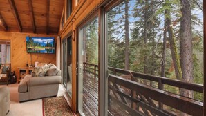 A cozy cabin interior with a large sliding glass door leading to a deck with a wooden railing, offering a view of tall trees. - Lake Tahoe Treasure! Minutes From Beach & Skiing (Homewood)