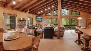 A spacious cabin interior with a dining area, seating, a fireplace, and large windows offering views of the outdoors. - Lake Tahoe Treasure! Minutes From Beach & Skiing (Homewood)