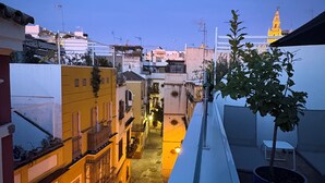 Une rue étroite bordée de bâtiments des deux côtés, un balcon orné d’un pot de fleurs et, au loin, une tour illuminée.