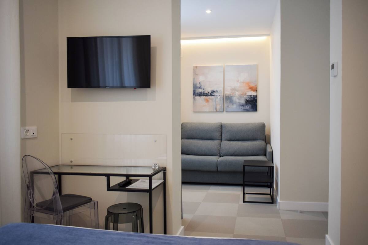 Um espaço de estar moderno e compacto, com televisão de tela plana na parede, sofá cinza, mesa pequena com uma cadeira e duas pinturas abstratas nas paredes.