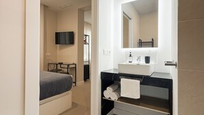 Une salle de bain moderne avec un grand miroir, un lavabo et des toilettes. Une chambre adjacente contient un lit.