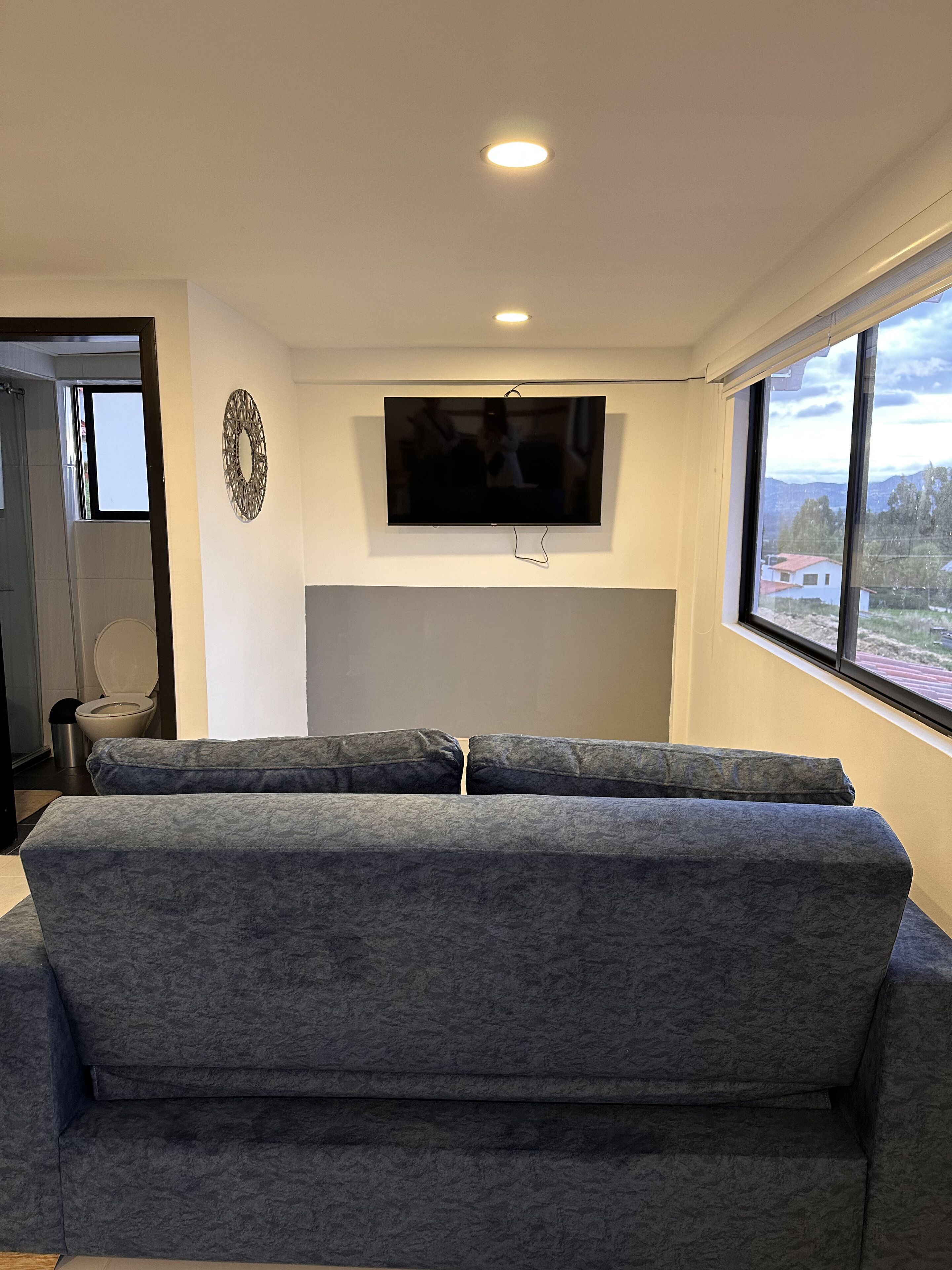 Un salon moderne avec un canapé gris, un téléviseur à écran plat fixé au mur et une grande fenêtre donnant sur l’extérieur.